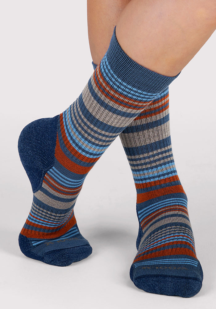 A person models Woolx Womens Everyday Crew Socks in Majolica Blue Stripe, featuring blue, gray, orange, and white stripes, with one foot slightly raised against a plain light background.