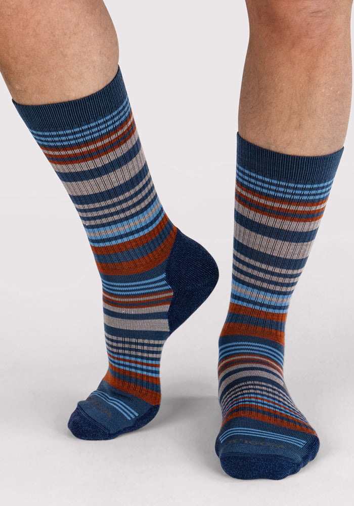 A person wearing Woolx Mens Everyday Crew Socks in Majolica Blue Stripe—colorful, moisture-wicking socks with blue, orange, gray, and black horizontal stripes—shown on lower legs and feet against a plain light background.