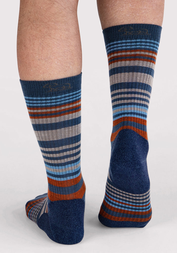 A person is wearing Woolx Mens Everyday Crew Socks in Majolica Blue Stripe, featuring horizontal blue, gray, orange, and white stripes with a darker heel and toe on a light gray background for a stylish look.