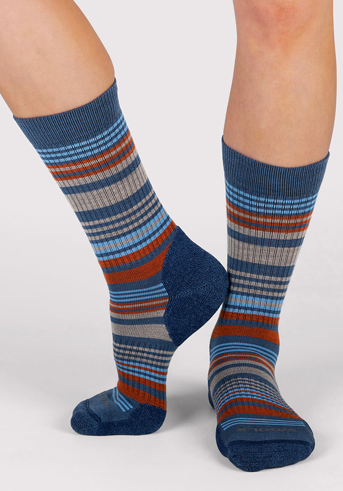 Two feet in Woolx Womens Everyday Crew Socks - Majolica Blue Stripe stand on a light gray surface, with one foot flat and the other raised on its toes.
