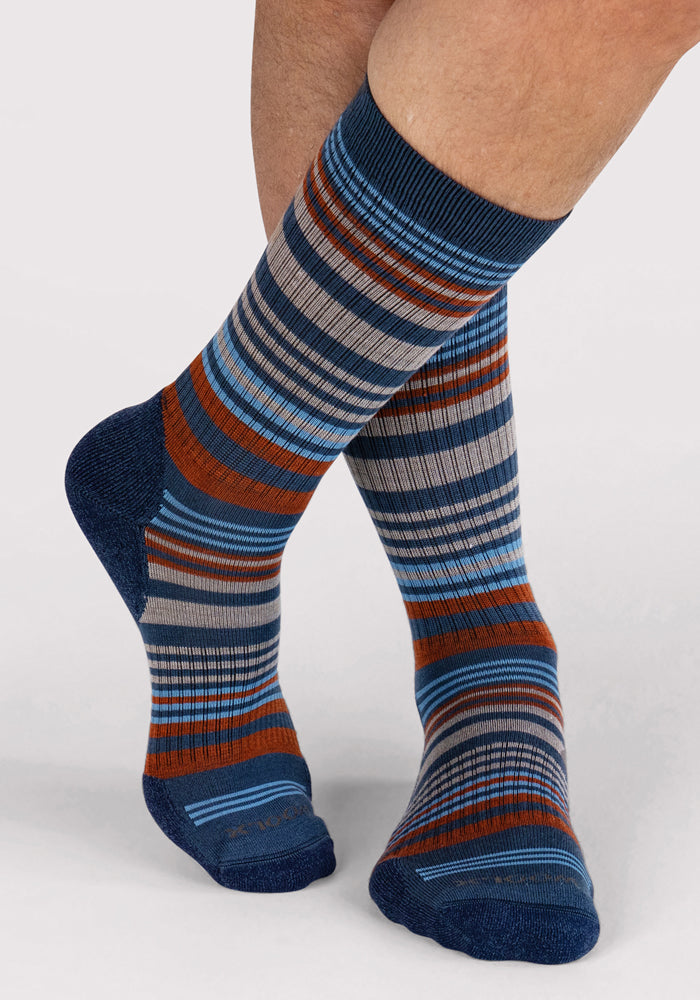 A person stands with one leg crossed over the other, wearing Woolx Mens Everyday Crew Socks in Majolica Blue Stripe with horizontal stripes in blue, brown, and gray against a plain background.
