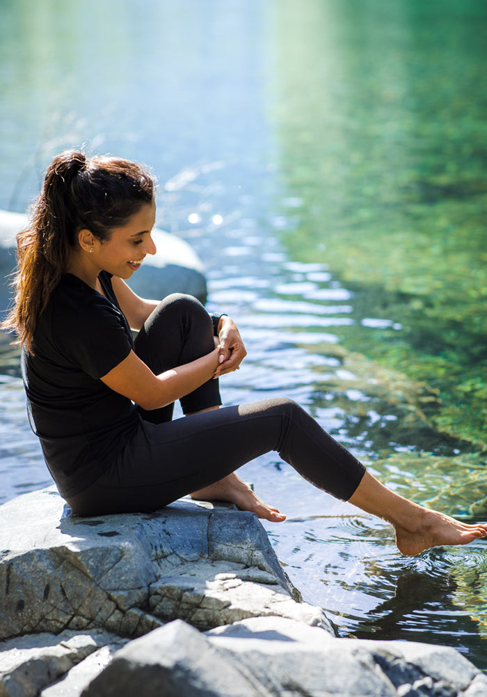 A woman wearing Woolx Merino Flex® McKenna Capris in black sits on a rock by clear water, smiling as she dips her bare feet into the stream on a sunny day.