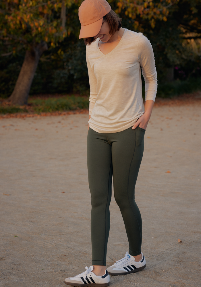A person in a light long-sleeve shirt, Woolx Merino Flex® McKenna Pocket Leggings in Everleaf, white Adidas sneakers, and a peach baseball cap stands on a dirt path outdoors with trees behind them, hands in pockets and looking down.