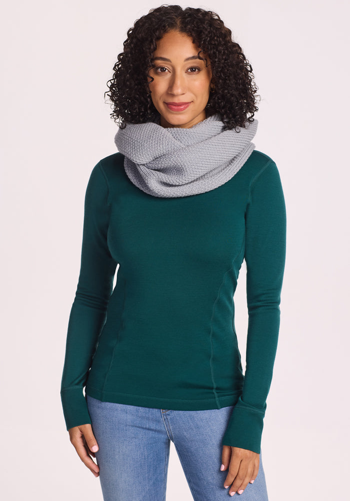 A woman with curly hair wears a dark green long-sleeve top, light blue jeans, and the Woolx Noelle Infinity Scarf in Morning Fog, standing against a plain light background and looking at the camera with a slight smile.