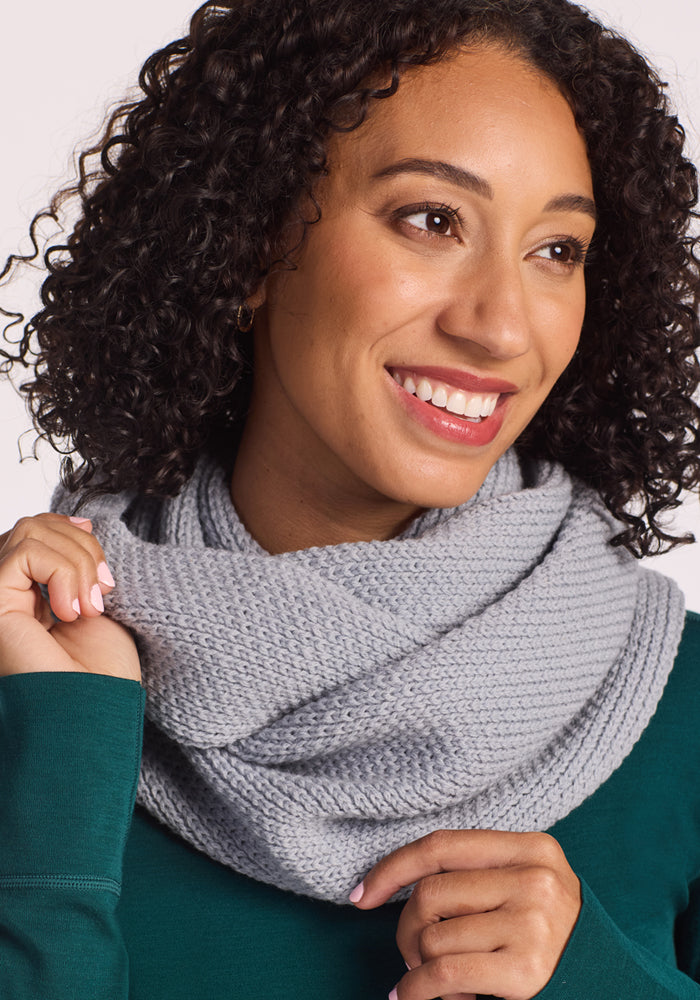 A woman with curly hair in a green top smiles, looking slightly to the side, as she holds part of the cozy Woolx Noelle Infinity Scarf - Morning Fog.