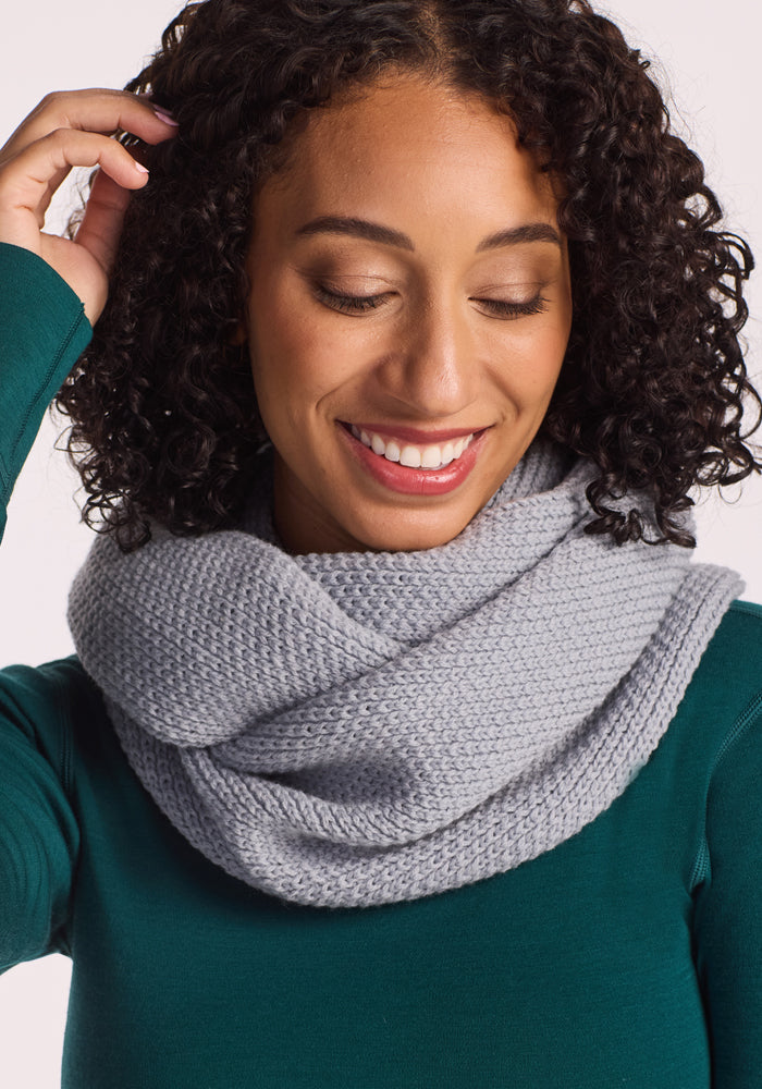 A smiling woman with curly hair looks down, touching her hair while wearing a teal top and the Woolx Noelle Infinity Scarf - Morning Fog, wrapped cozily around her neck.