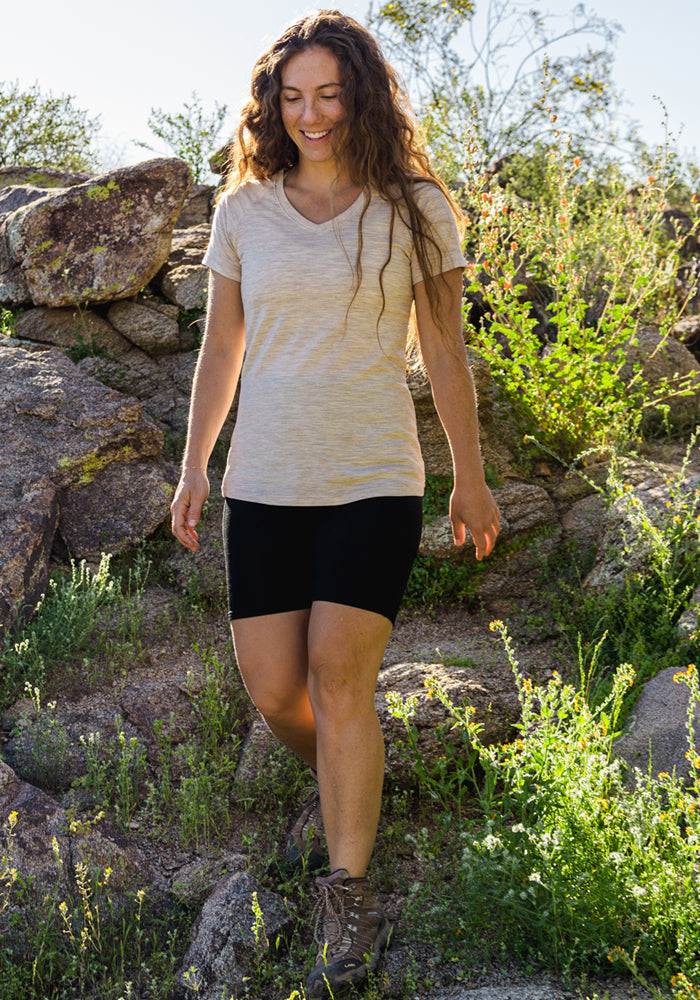 A woman with long wavy hair wears the Woolx Mia V Neck in Cream Heather, paired with black shorts and hiking boots, as she walks outdoors on rocky terrain surrounded by greenery and sunlight.