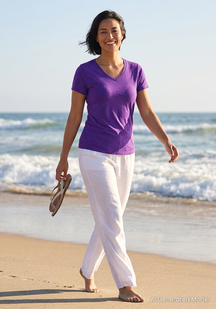 A woman with short dark hair walks barefoot on a sandy beach, holding sandals and wearing the Woolx Mia V Neck - Plum Crush moisture-wicking t-shirt and white pants, with gentle waves and a clear sky behind her.