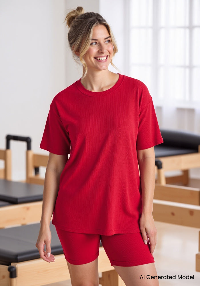A smiling woman wears the Woolx Vera Ribbed Tunic in Poppy Spark, standing in a bright room with Pilates reformers.