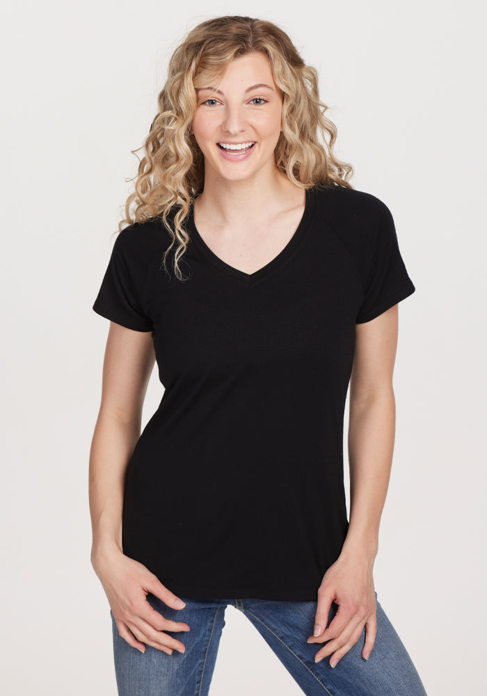A person with curly blonde hair is smiling while wearing the women's Mia V Neck in Black by Woolx, crafted from premium merino wool, paired with blue jeans. They are standing against a plain white background. | Jordan is 5’6”, wearing a size S