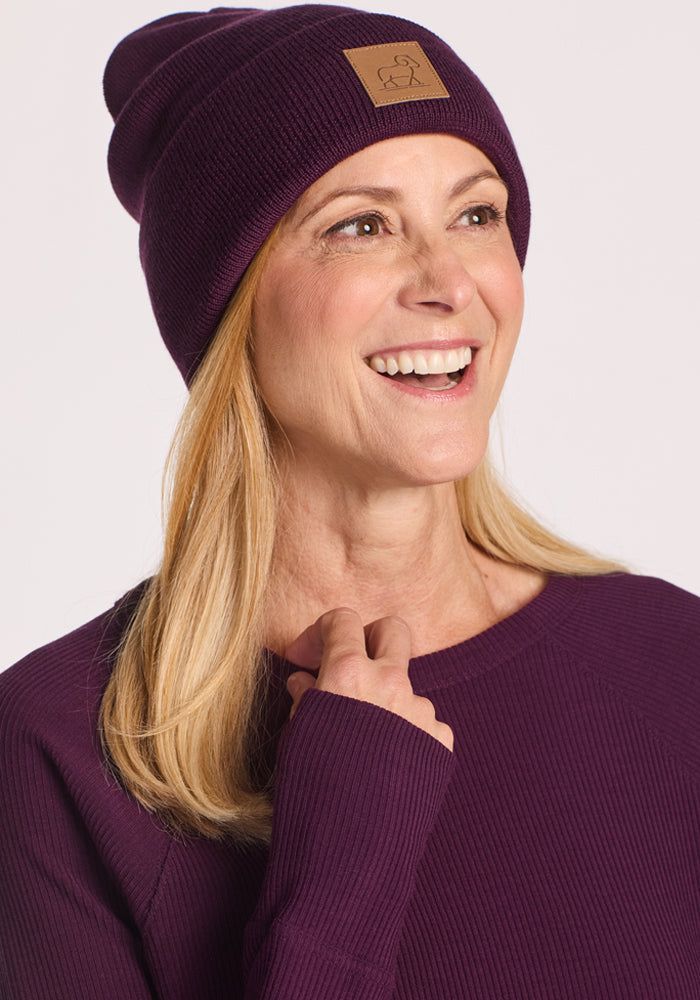 A smiling woman with blonde hair wears the Woolx Baylor Beanie in Majestic Purple, paired with a matching ribbed sweater. She looks off to the side against a light background, radiating cozy winter style in this warm hat.