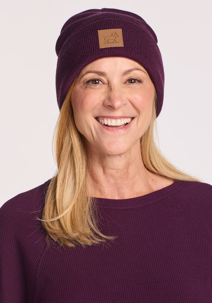 A smiling woman with long blonde hair wears the Woolx Baylor Beanie in Majestic Purple, featuring a brown patch with a simple line design. Her matching sweater and the plain light background highlight this cozy winter hat.