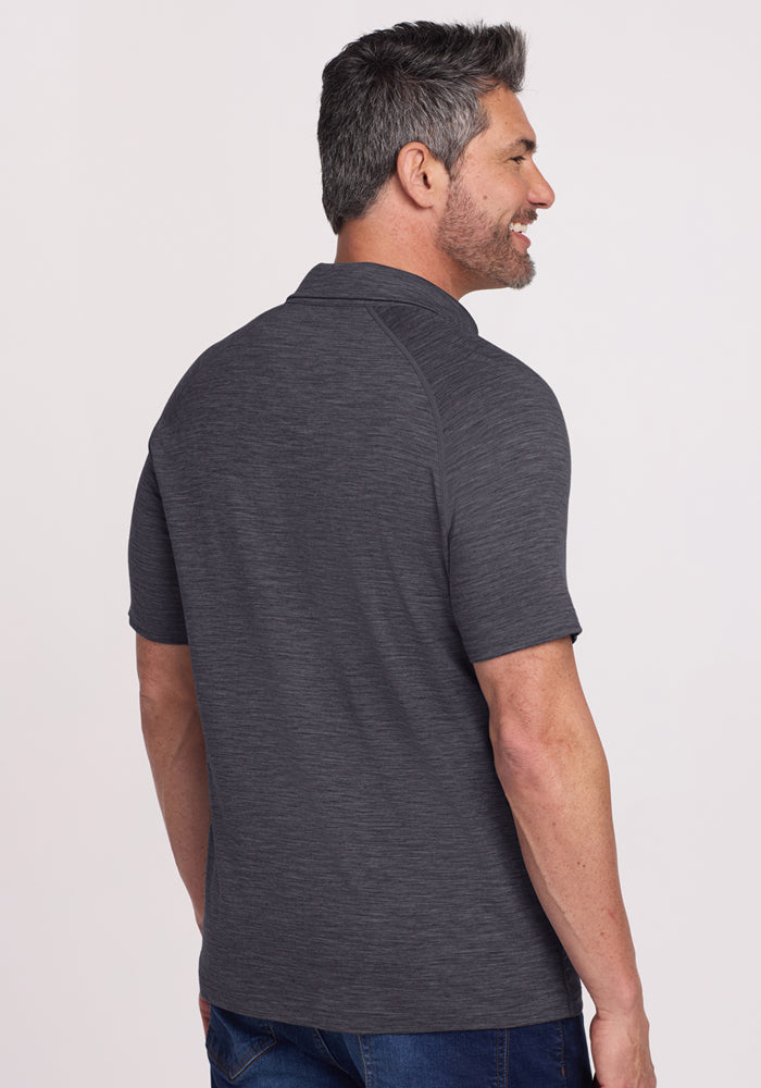 A man with short, gray hair and a beard stands facing away from the camera in blue jeans and the Woolx Summit Polo - Cinder Melange against a plain light background.