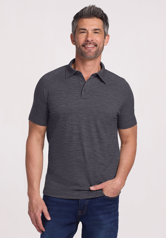 A middle-aged man with short dark hair and a trimmed beard smiles while wearing the Woolx Summit Polo in Cinder Melange and blue jeans, standing against a light background, showcasing effortless style and moisture management. 