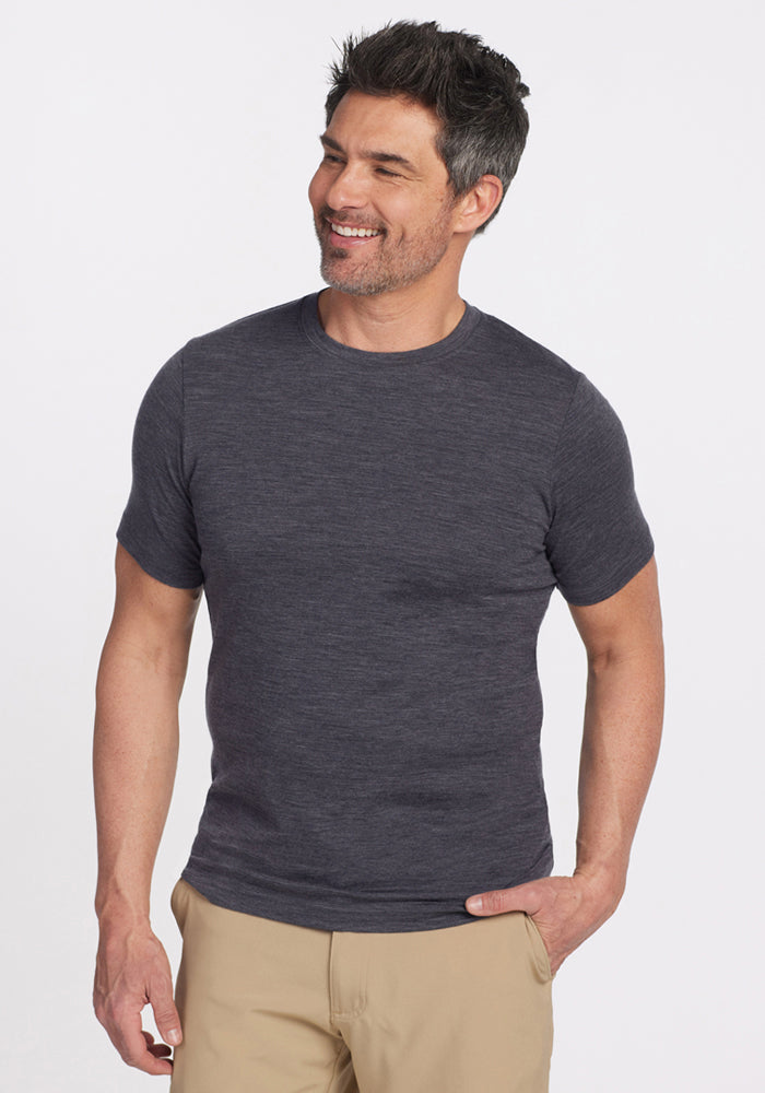 A man with short dark hair and light facial hair smiles while wearing the Woolx Endurance Tee in Cinder Melange and beige pants, standing against a plain white background. 