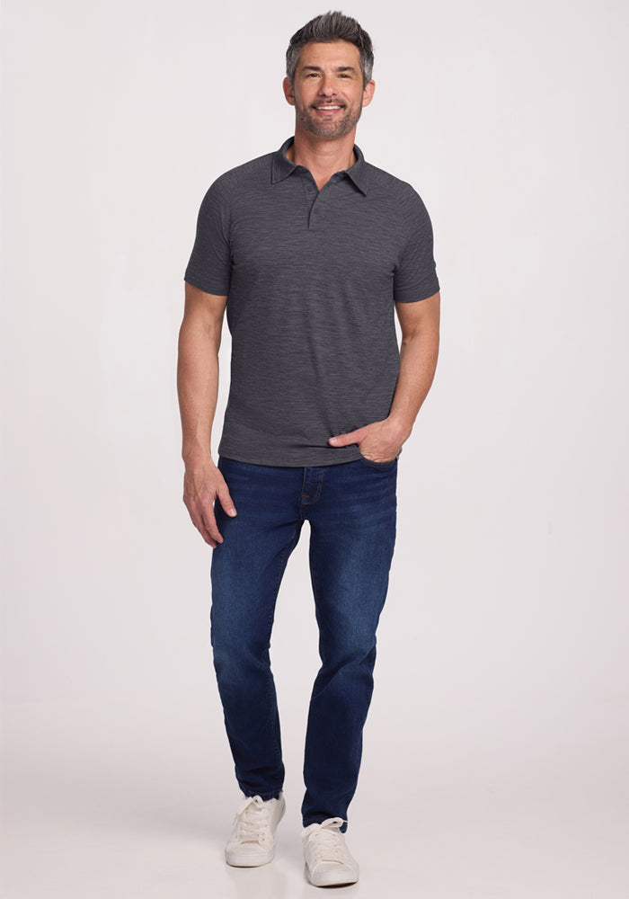 A man with short dark hair and a beard smiles while standing against a plain light background, wearing the Woolx Summit Polo - Cinder Melange, blue jeans, and white sneakers, with one hand in his pocket.