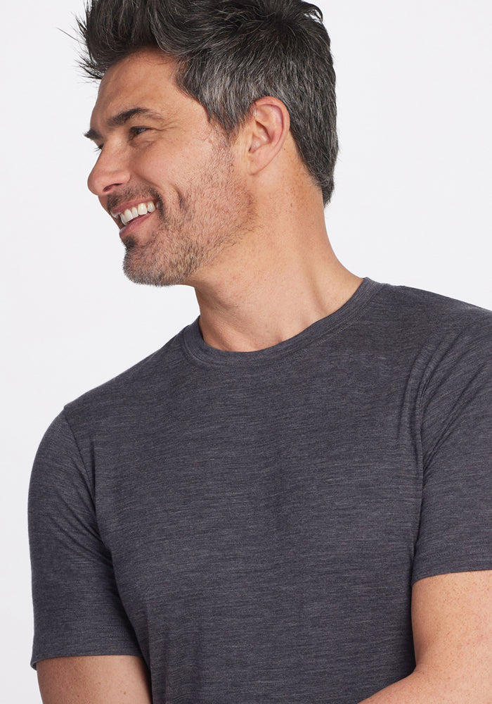 A man with short, salt-and-pepper hair and a beard is smiling and looking to his left. He is wearing the Woolx Endurance Tee in Cinder Melange against a plain white background.