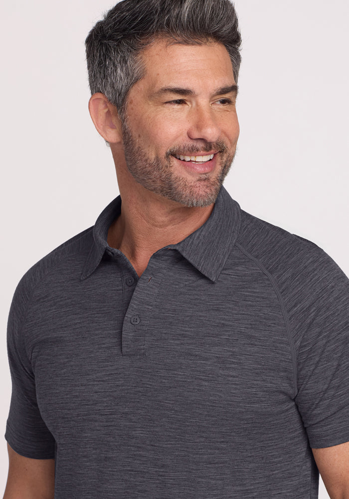 A man with short salt-and-pepper hair and a beard smiles, looking to the side. He wears the Woolx Summit Polo in Cinder Melange, made from Merino wool for excellent moisture management. The background is plain and light-colored.