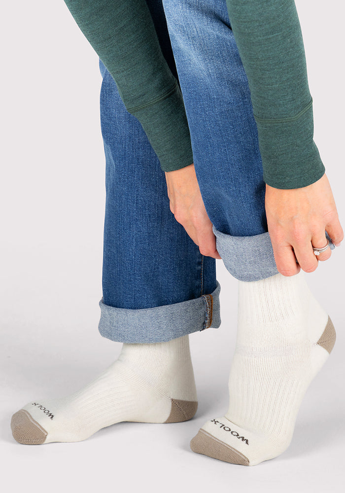 A person in blue jeans and a green long-sleeve shirt rolls up their pant leg, revealing Woolx Womens Core Crew Socks - Natural, featuring cushioned beige toes and heels with "WOOL" visible near the toe.