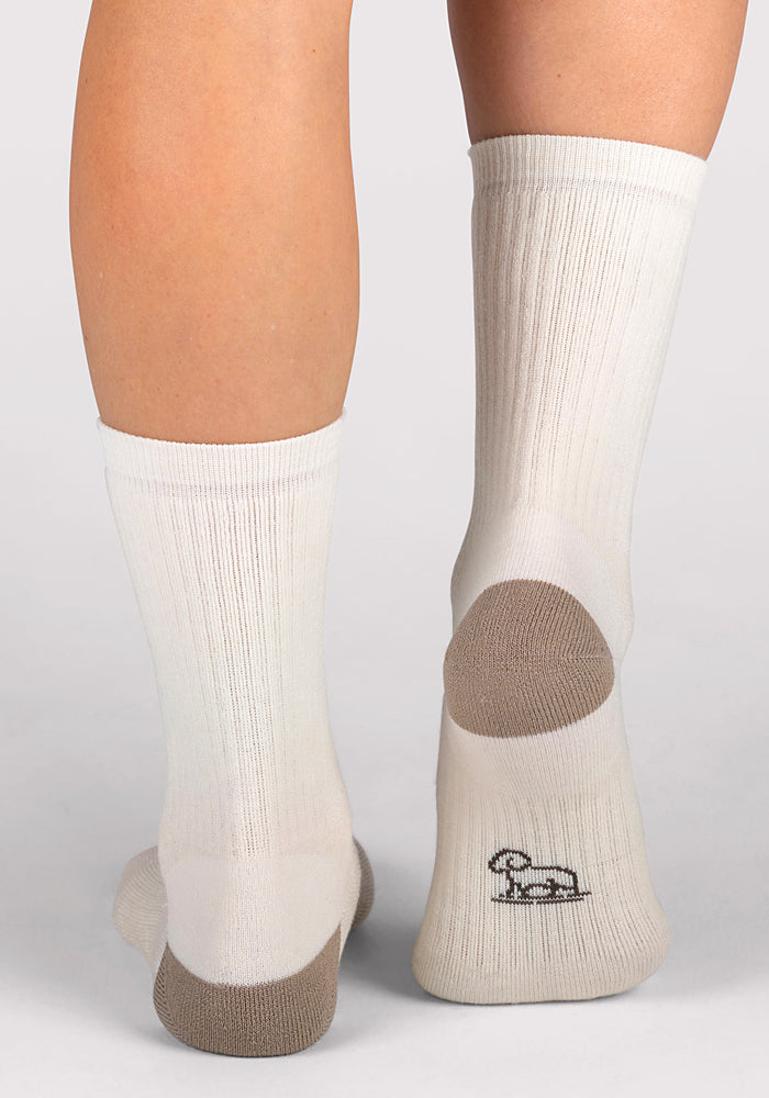 A person wearing Woolx Womens Core Crew Socks - Natural with moisture-wicking fabric and gray heels and toes stands on a light surface. A small black sheep logo is visible on the bottom of the right sock.