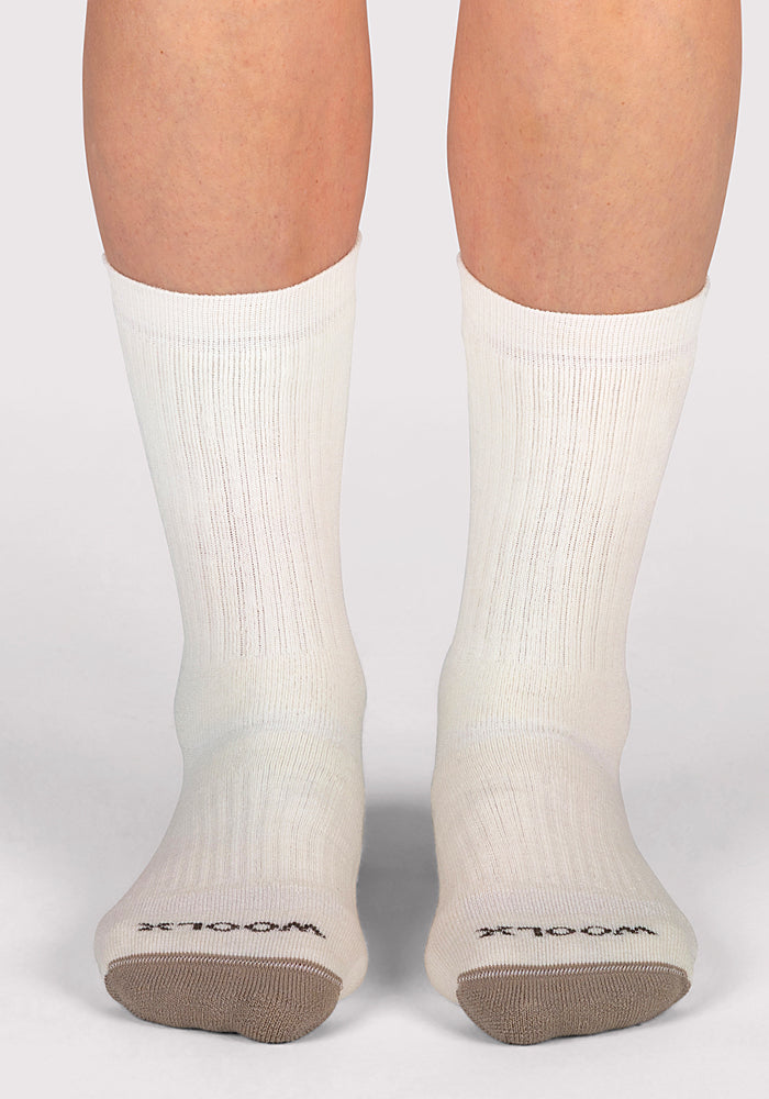 A pair of feet wear Woolx Womens Core Crew Socks - Natural, featuring white cushioned fabric with beige toes and "WOOL" printed in black on top, standing on a light-colored surface.