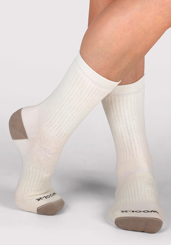 A pair of feet wears Woolx Womens Core Crew Socks - Natural, featuring a ribbed texture, tan heels and toes, moisture-wicking white fabric, and "WOOLX" on the soles, against a plain light background.