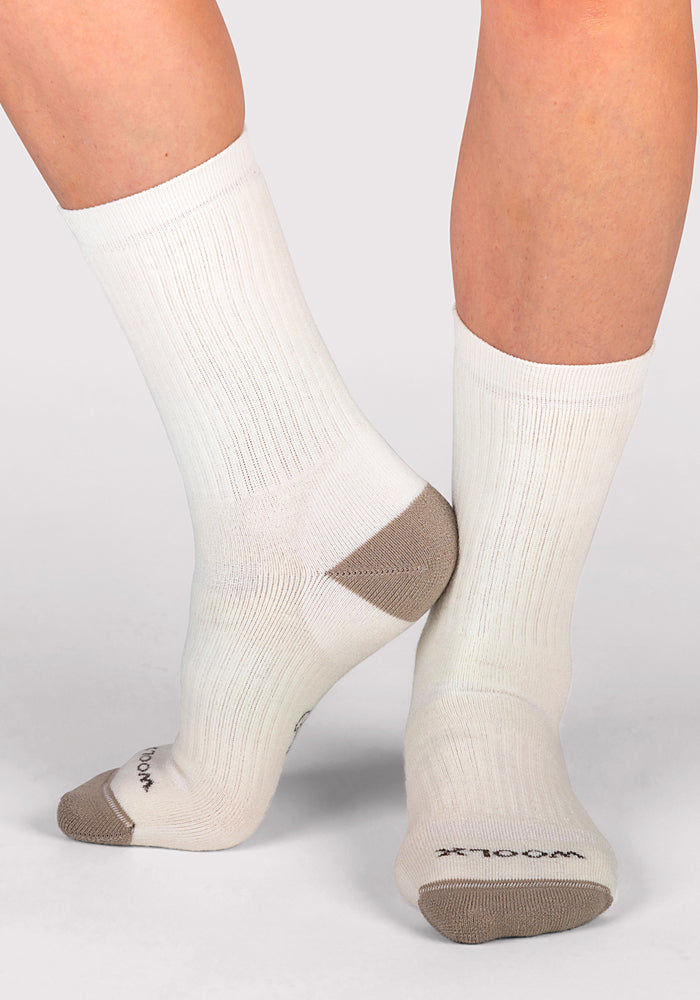 A person stands on a light gray surface wearing Woolx Womens Core Crew Socks - Natural, featuring white cushioning, beige heels and toes, and the brand name “Woolx” near the toes. The socks are moisture-wicking.