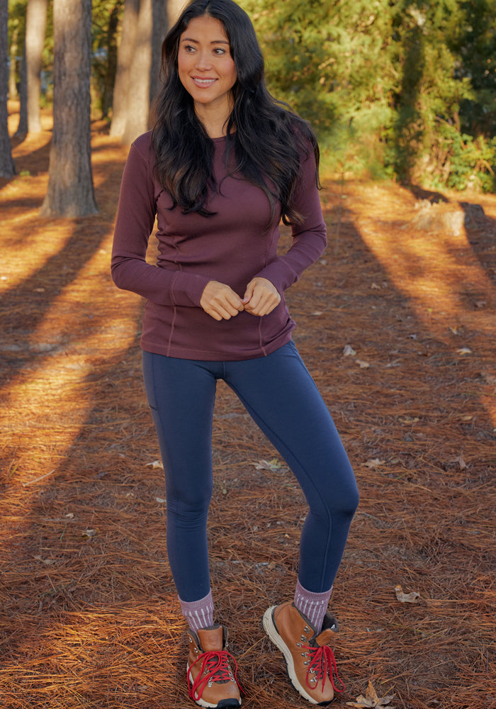 A woman stands outdoors on pine needles, smiling in a maroon top and Woolx Piper Pocket Leggings in Toasted Garnet. She wears moisture-wicking hiking shoes with thick socks, surrounded by sunlit tall trees.