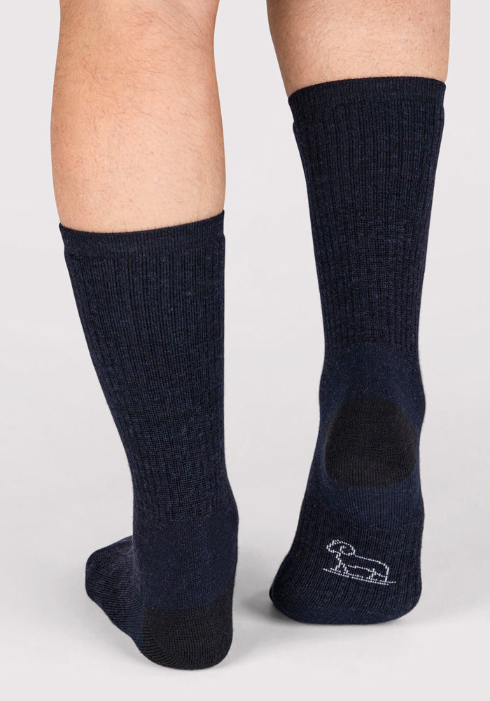 A close-up shows a person's lower legs wearing Woolx Mens Core Crew Sock in navy, featuring dark blue, moisture-wicking ribbed fabric and a small white dog outline near the heel against a plain light background.