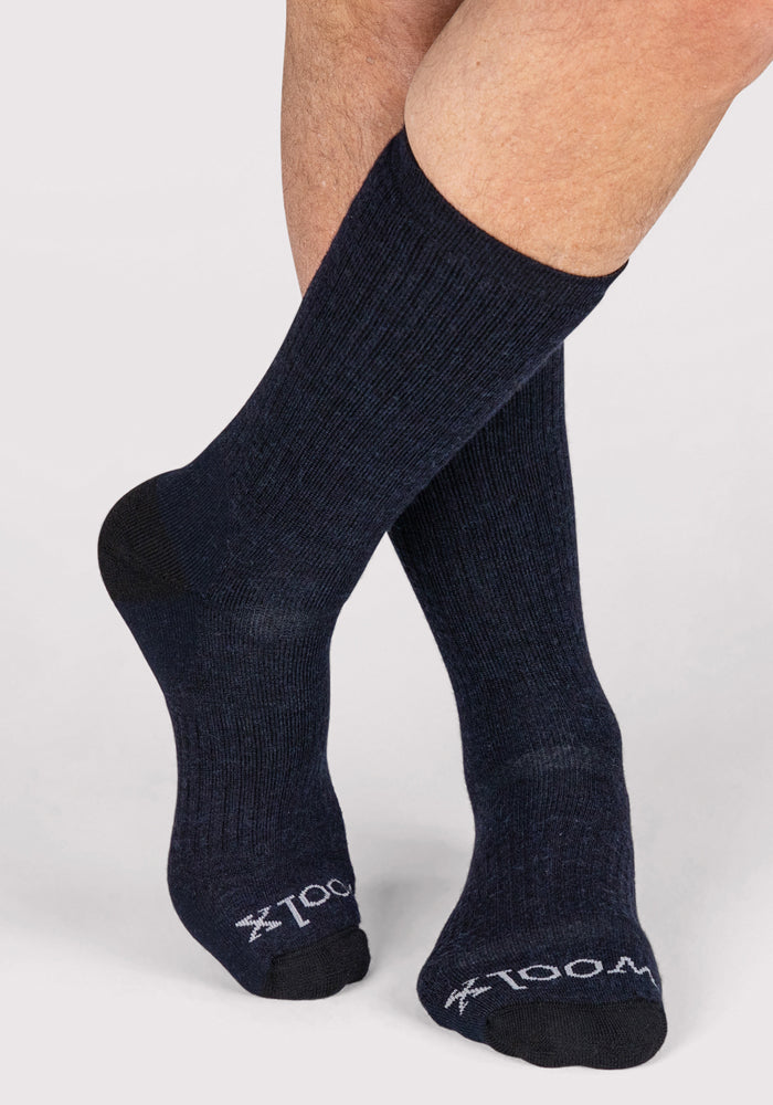 A person wearing Woolx Mens Core Crew Sock in navy with black toes and heels. The Merino wool socks feature white text and a logo near the toes, and their legs are crossed at the ankles against a light background.