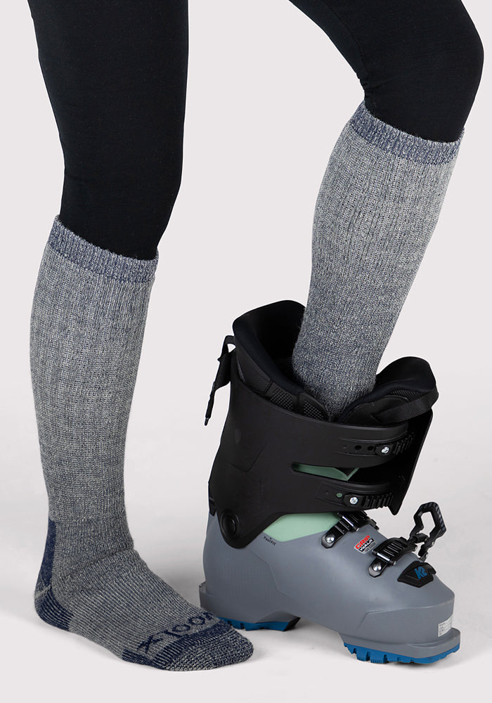 A person wearing Woolx Womens Extreme Over The Calf Socks - Navy Carbon places one foot into a gray and black ski boot while the other foot, in just the sock, stays cozy—ideal for warmth in cold weather.