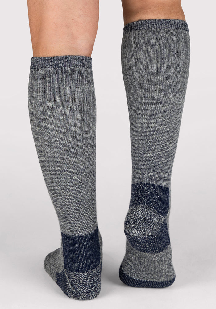 A person wearing Woolx Mens Extreme Over The Calf Socks in Navy Carbon, featuring dark blue accents on the heel, toe, and sole, stands on a plain white background with the backs of their lower legs and feet visible.