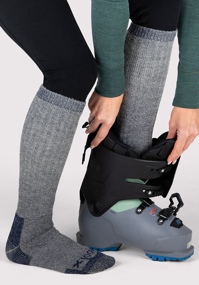 A person wearing Woolx Womens Extreme Over The Calf Socks in Navy Carbon and black leggings is putting on a gray ski boot, with one foot already inside and hands adjusting the boot closures.