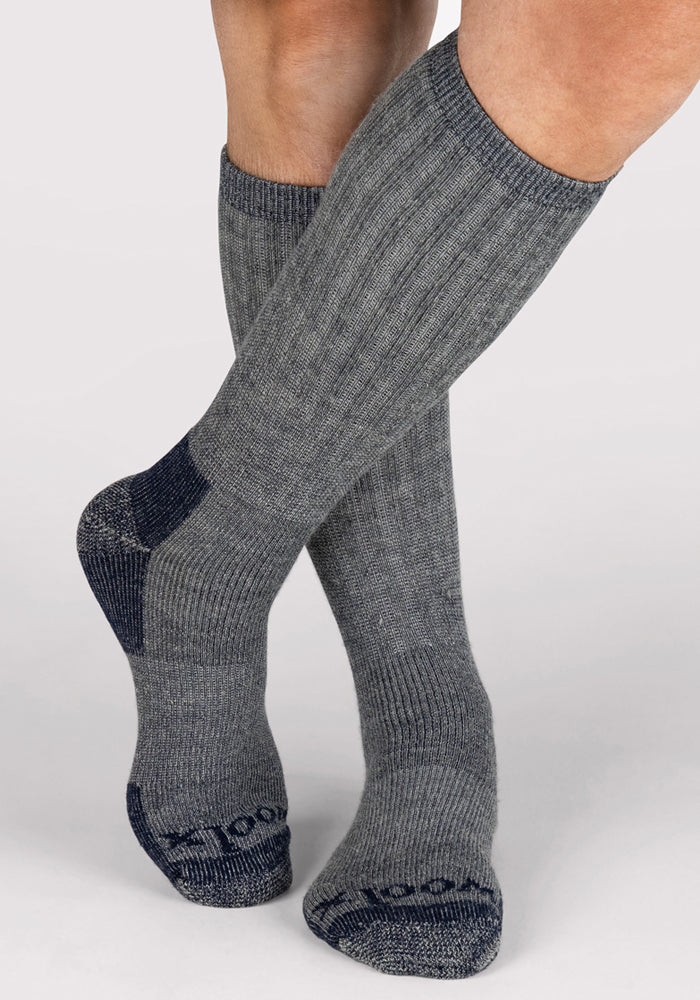 A person crosses their bare legs, wearing Woolx Mens Extreme Over The Calf Socks in Navy Carbon, featuring ribbed navy fabric with carbon accents on toes, heels, and soles. The plain backdrop evokes cozy cold-weather moments indoors.