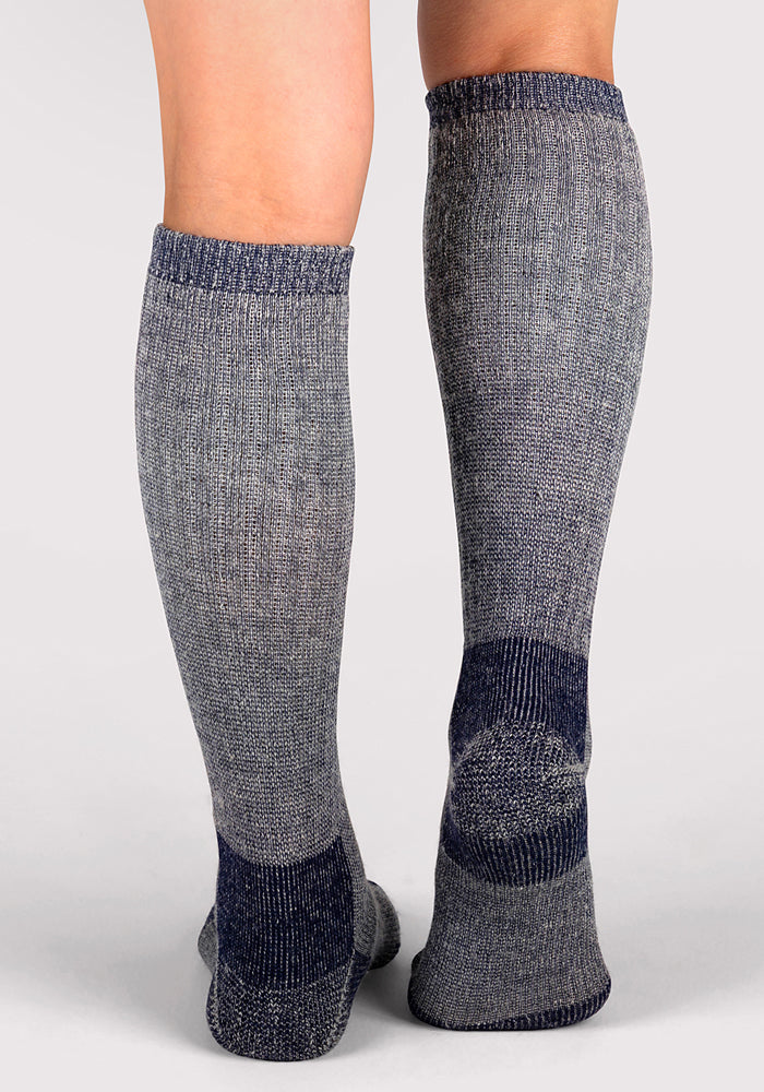 A person stands on a light surface, wearing Woolx Womens Extreme Over The Calf Socks in Navy Carbon—heathered gray with darker blue reinforced heels, toes, and cuffs. The heavyweight Merino wool socks are ideal for cold weather, shown from behind.