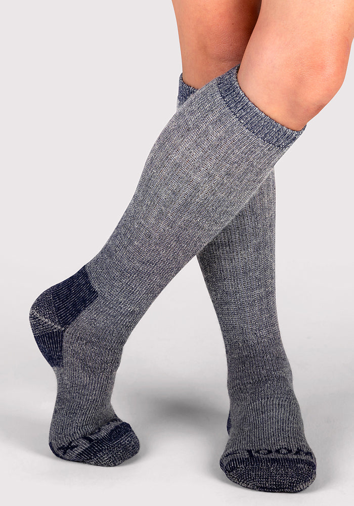 A person wears Woolx Womens Extreme Over The Calf Socks in Navy Carbon, featuring dark gray accents on the toes, heels, and cuffs, standing with one leg crossed in front of the other against a light background.