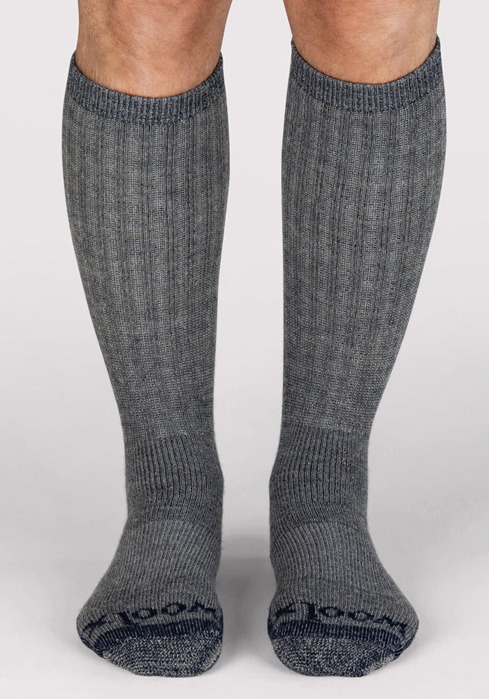 A person wears Woolx Mens Extreme Over The Calf Socks in Navy Carbon with a ribbed texture, standing on a light background. Only the lower legs and feet are visible, ideal for cold-weather activities.
