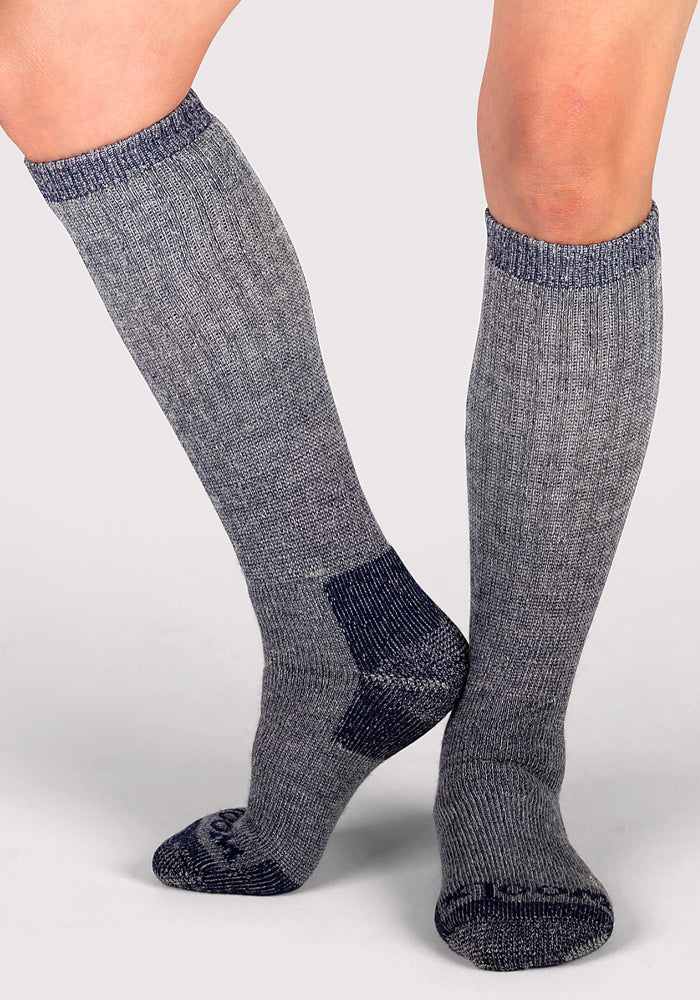 A person wears Woolx Womens Extreme Over The Calf Socks in Navy Carbon, featuring heavyweight Merino wool, with darker heel, toe, and cuff accents, standing against a plain light background.