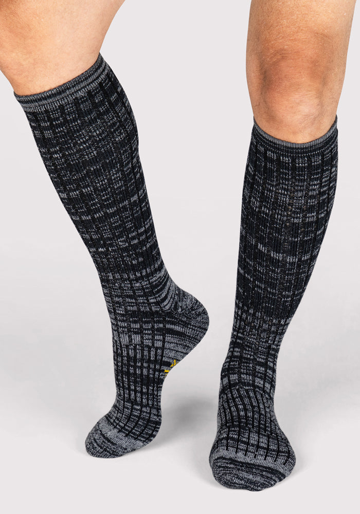 A person wearing Woolx Men's Joey Mid Calf Socks in Dark Navy Marled stands on a light gray background, one foot slightly raised and toes pointed down.
