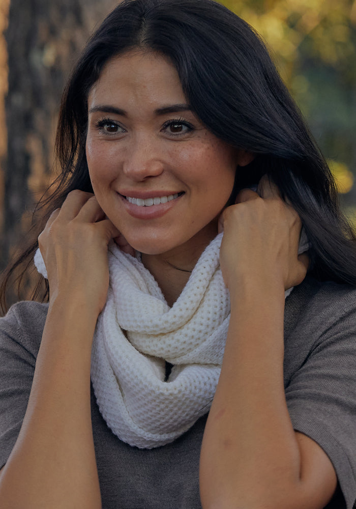 A woman with long dark hair smiles while wearing the Woolx Noelle Infinity Scarf in Buttercream, wrapped around her neck over a grey top. She stands outdoors with blurred trees, highlighting this cozy winter accessory.