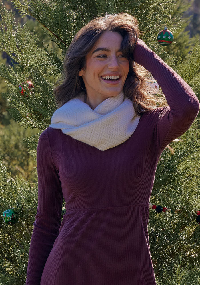 A woman wearing the Woolx Noelle Infinity Scarf in Buttercream smiles with one hand in her hair, standing outdoors before a decorated evergreen tree.