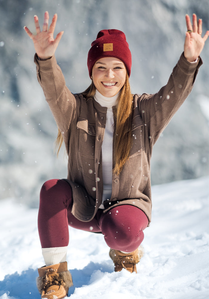A woman kneels in the snow, wrapped in the Woolx Oaklynn Waffle Shacket in Latte.