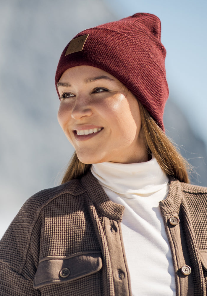 A smiling woman in a burgundy knit beanie, white turtleneck, and the Woolx Oaklynn Waffle Shacket in Latte stands outdoors against a snowy, blurred background.