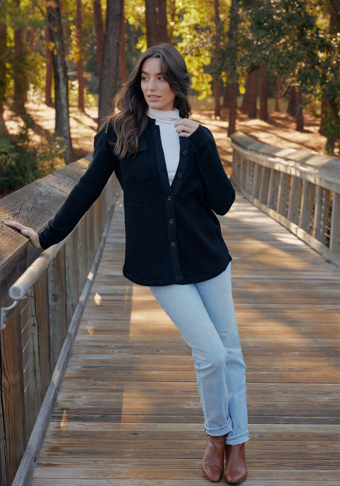 Wearing the Woolx Oaklynn Waffle Shacket in Latte over a white turtleneck, light blue jeans, and brown boots, a woman stands on a wooden bridge in a forested area, leaning on the railing as sunlight filters through the trees.