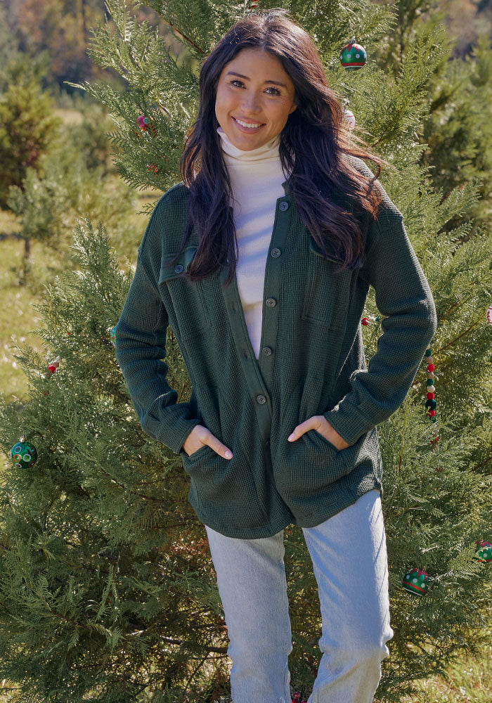 A woman with long dark hair stands in front of a decorated outdoor evergreen, smiling with her hands in her pockets, wearing the Woolx Oaklynn Waffle Shacket in Driftwood Fern over a white turtleneck and light blue jeans.