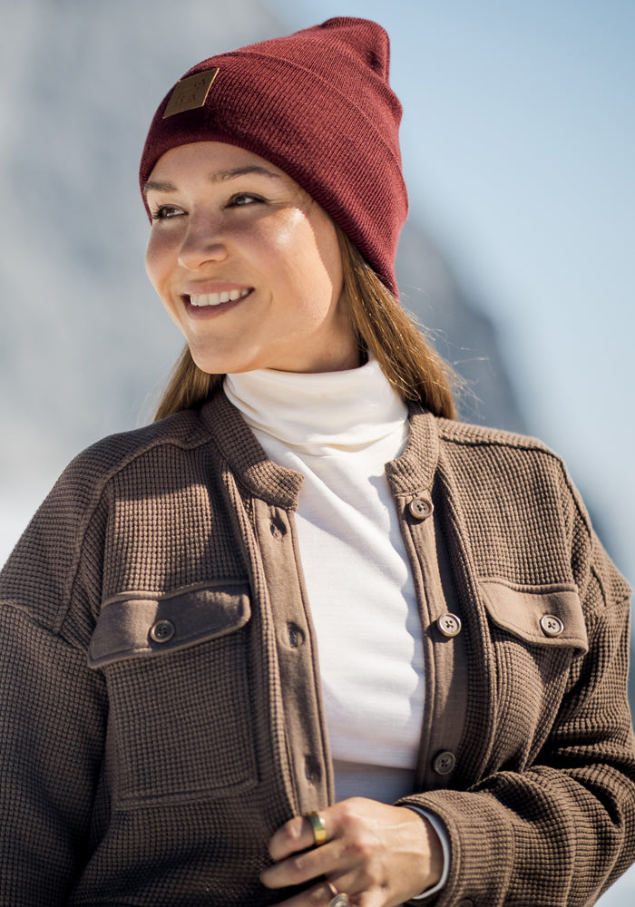 A smiling woman in a Woolx Oaklynn Waffle Shacket - Latte, a burgundy beanie, and a white turtleneck stands outdoors with a blurred snowy background.