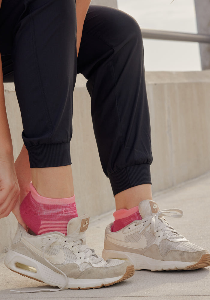 A person adjusts their bright pink ankle socks while wearing white Nike sneakers and Woolx Palmer Joggers on concrete steps. Only the lower legs and feet are visible.