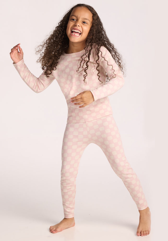 A joyful young girl with long curly hair smiles while wearing Woolx Kids Explorer Base Layer Bottoms in Hopscotch Pink. She stands barefoot against a plain light background, looking ready for adventure.