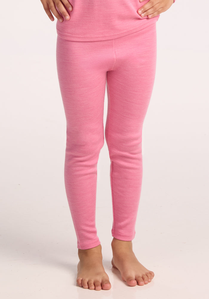 A child stands barefoot in Woolx Kids Explorer Base Layer Bottoms - Pinky Promise, hands on hips, shown from shoulders down against a plain light background.