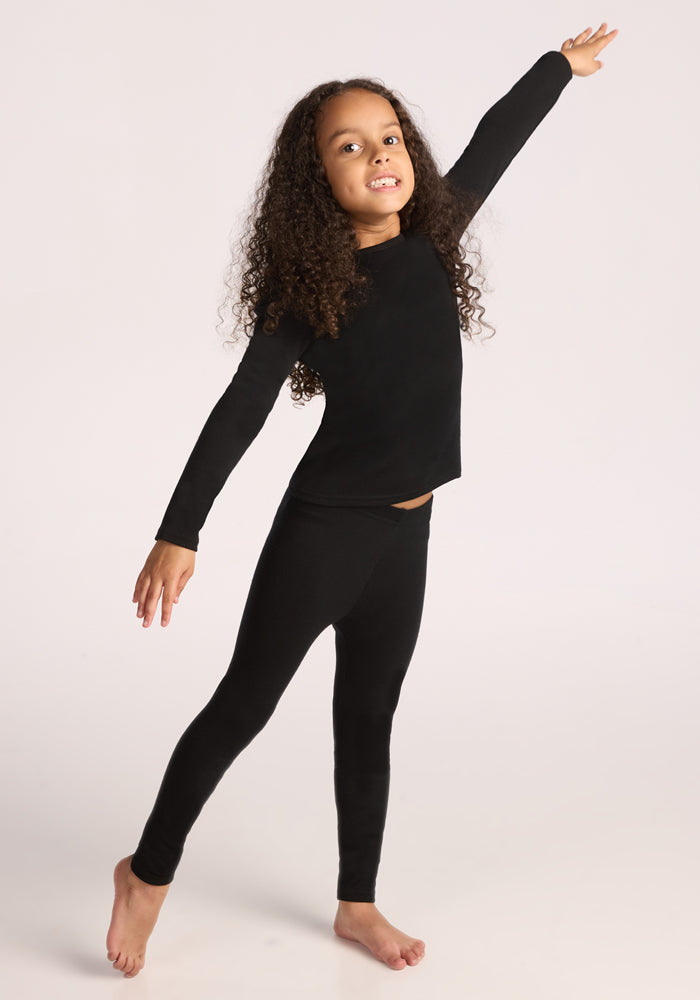 A young child stands barefoot on one foot with arms outstretched, smiling in Woolx Kids Explorer Base Layer Bottoms - Black against a light background.